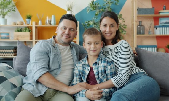 Portrait of happy young family mother, father and son sitting on sofa at home smiling looking at camera. People, lifestyle and togetherness concept.